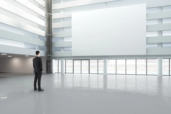 Uomo guardando un banner bianco vuoto in una grande sala luminosa, beffa — Foto Stock