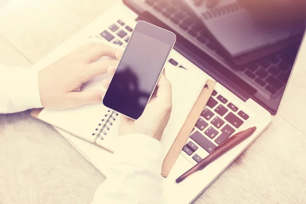 Mano femenina con un teléfono celular en blanco y un portátil sobre la mesa — Foto de Stock