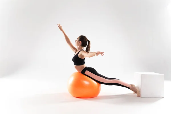 A slender girl in leggings and a sports top lies on a fitness ball on a white isolated background. Athlete girl on a white isolated background, with a fitness ball.