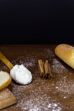 Tahta bir kaşıkta un, ahşap bir tahtanın üzerinde dilimlenmiş Fransız ekmeği ve ahşap bir masada siyah bir arka planı olan un unlu tarçın çubukları. Metin ya da logo için yer olan kenar görünümü. Retro fotoğraf.