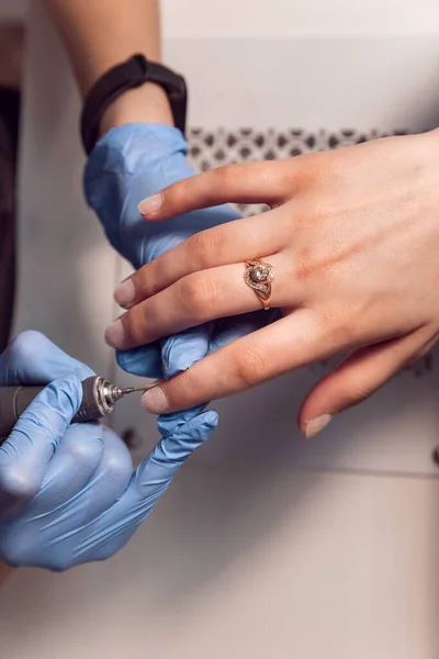 Manicure master doing manicure in blue gloves for client with a Electric Nail File Drill Machine. Female hardware manicure in the salon. Electric nail file for trimming and removing cuticles in beauty salon.