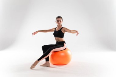 A slender girl in leggings and a sports top, stands on a white background, putting one leg on a fitness ball. Girl athlete on a white isolated background, with a ball