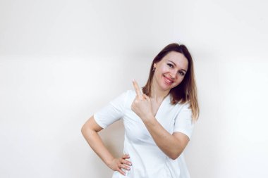 Güzellik uzmanı kameraya gülümser ve mesaj veya logo için parmağını yana doğru tutar. İzole edilmiş beyaz bir arka planda beyaz bir önlük içinde güzel bir bayan doktor..
