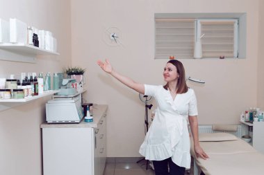 Beyaz cüppeli bir güzellik uzmanı güzellik salonunda duruyor ve ürünü eliyle işaret ediyor. Beyaz tıbbi önlüklü bir kozmetolog, ofisin arka planında, müşterileri kabul etmeye hazırlanıyor.