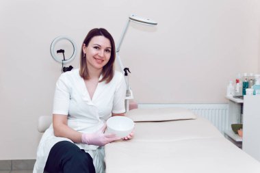 Cosmetologist doctor in a white medical coat, against the background of the office, prepares to receive clients. A cosmetologist in a white coat smiles and holds a white bowl in his hands.