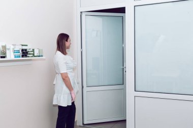 A cosmetologist in a white medical coat opened the door to wait for the client. A doctor in a white coat is waiting for a client.