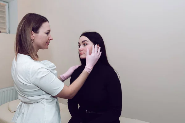 A cosmetologist in a white medical coat gets to know the client and makes a skin diagnosis. A doctor in a white coat checks the client's face. Cosmetologist with patient on medical chair.