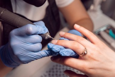Manikür ustası, elektrikli çivi dosya matkap makinesi olan bir müşteri için mavi eldivenli manikür yapıyor. Kuaförde kadın manikürü. Güzellik salonundaki tırnak etlerini budamak ve sökmek için elektrikli tırnak törpüsü.