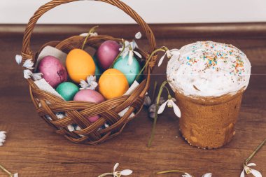 Easter background. Easter eggs in a basket and Easter pastries. Kulich. On a wooden table is an Easter basket with yellow, green and purple eggs, decorated with snowdrops.