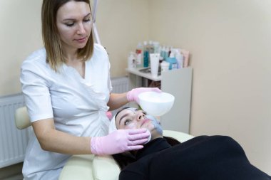 Kozmetik maske takan bir kadın. Beyaz tıbbi elbise giyen güzellik uzmanı müşterinin yüzündeki kozmetik maskeyi yıkar. Güzellik salonunda kozmetik maskeli güzel bir kadın..