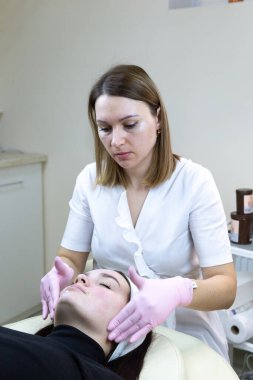 Kozmetik maske takan bir kadın. Beyaz elbiseli bir güzellik uzmanı güzellik salonundaki güzel bir kadına krem sürüyor..