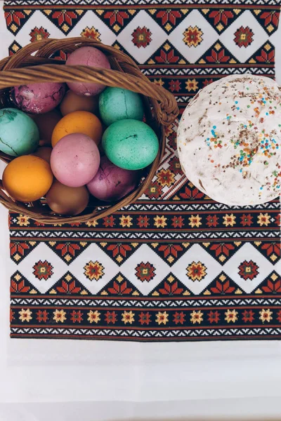 Easter eggs and Easter cake on an embroidered towel. Ukrainian embroidered shirt and Easter cake on a wooden table with Easter eggs of different colors - yellow eggs, green and purple eggs in a basket.