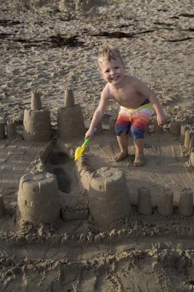 Vacker ung blond pojke leker på stranden i ett sandslott — Stockfoto