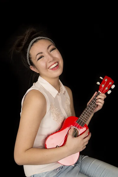 Beautiful older teen girl playing a red ukulele sitting smiling  black background — Stock Photo, Image