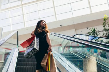 Markette yürürken renkli alışveriş torbaları taşıyan şık siyah elbiseli neşeli bir bayan. Çekici esmer, alışveriş merkezinden alışveriş yaptıktan sonra tatmin olmuş görünüyor..