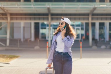 Genç ve güzel bir kız, elinde bavulla büyük bir otobüs durağında telefonla konuşuyor.