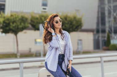 Genç ve güzel bir kız, taksi bekliyor, ve büyük kulaklıklarla telefondan müzik dinliyor.