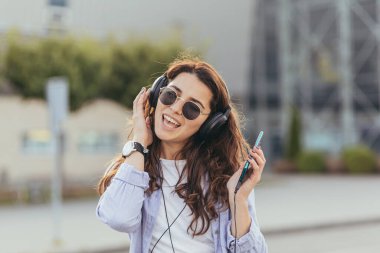 Genç ve güzel bir kız, taksi bekliyor, ve büyük kulaklıklarla telefondan müzik dinliyor.