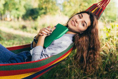 Genç ve güzel bir kız parktaki renkli bir hamakta dinleniyor ve kitap okuyor.
