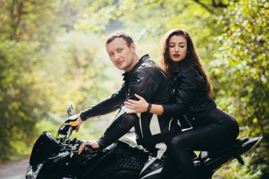 Genç bir çift, siyah spor bir motosikletin yanındaki adam ve kız, siyah deri elbiseli, ormanda yolda.