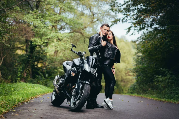 Genç bir çift, siyah spor bir motosikletin yanındaki adam ve kız, siyah deri elbiseli, ormanda yolda.
