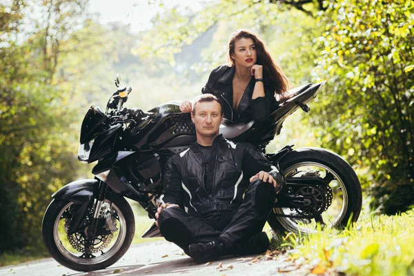 Genç bir çift, siyah spor bir motosikletin yanındaki adam ve kız, siyah deri elbiseli, ormanda yolda.