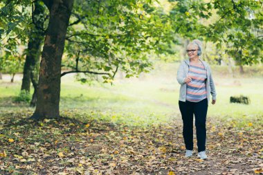 Sırt çantasıyla parkta yürüyüp kulaklıkla müzik dinleyen güzel, son sınıf öğrencisi bir kadın.