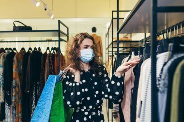 Yüzünde koruyucu maske olan genç ve güzel bir kadın mağazadan kıyafet seçiyor ve güvenli alışverişler yapıyor.