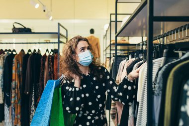 Yüzünde koruyucu maske olan genç ve güzel bir kadın mağazadan kıyafet seçiyor ve güvenli alışverişler yapıyor.