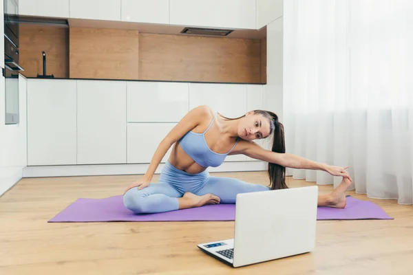 Beautiful fitness woman doing fitness exercises, looking for online tutorials on laptop, training in the kitchen. Healthy Lifestyle. The girl goes in for sports at home.