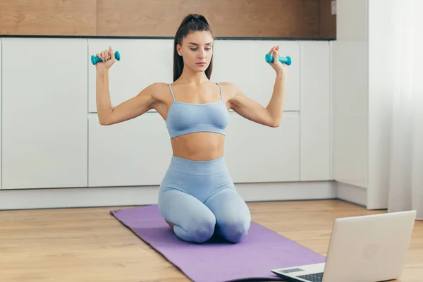 Beautiful fitness woman doing fitness exercises, looking for online tutorials on laptop, training in the kitchen. Healthy Lifestyle. The girl goes in for sports at home.
