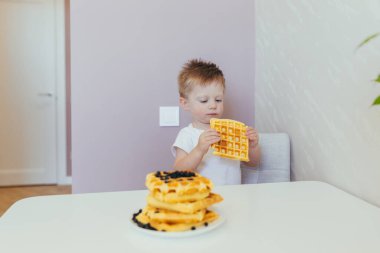 Beyaz elbiseli küçük bir çocuk mutfağında waffle ile kahvaltı yapıyor.