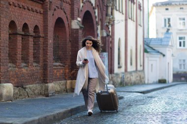 Yeni bir şehirde elinde telefon ve bavuluyla yürüyen güzel esmer bir kadın, bir turist yeni şehirler keşfediyor.