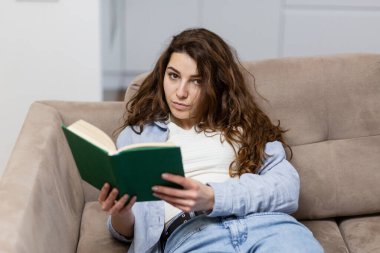 Güzel, genç bir kadın evinde kanepede oturup ilginç bir kitap okuyor.