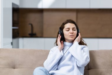 Genç ve güzel bir kadın evde dinleniyor, kanepede oturuyor ve kulaklıkla müzik dinliyor.