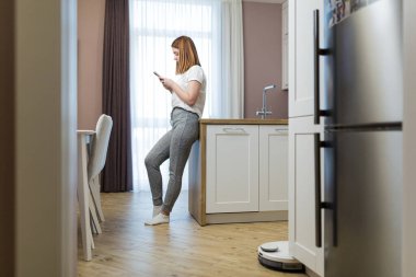 Genç ve güzel bir kadın ev işinden dinlenirken robot bir elektrik süpürgesi daireyi temizliyor.