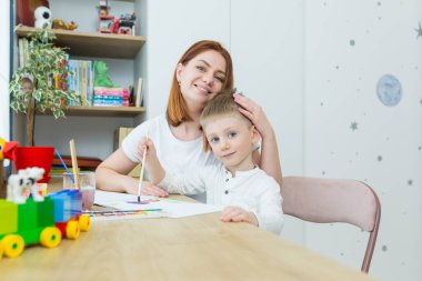 Genç anne, küçük oğluna resim çizmeyi öğretiyor, evde çocuk odasında, birlikte eğleniyor.
