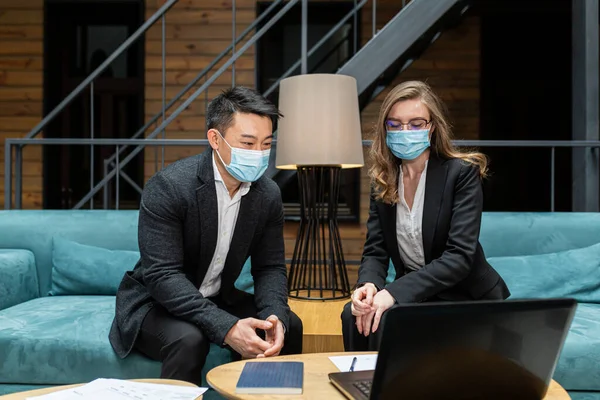 Two employees of an international company discuss a report on the work done, an Asian man and a woman wearing protective medical masks, sitting in the office at the computer