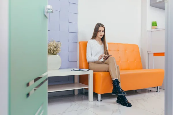 young woman in the waiting room interview sitting on sofa in corridor of dentist office or reception modern medical center while doctor\'s appointment. using mobile phone fills information or resumes