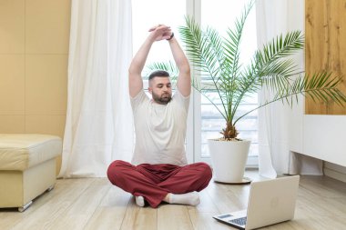 Genç, sakallı bir adam internette laptopla yoga yapıyor. Lotus pozisyonunda yerde oturan bir adam egzersiz yapıyor. Oturma odasında gerinen video derslerine bakıyor..