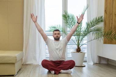 Genç sakallı yetişkin adam oturma odasında lotus pozisyonunda yerde oturmuş yoga yapıyor. Rahatlama ve meditasyon tek başına erkek nefes alma egzersizleriyle meşgul. Rahatla ve sabah dinlen.