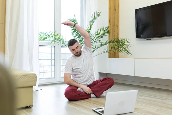 young adult bearded man doing stretching exercises at home in the living room watching video lesson or tutorial online using a laptop. Man doing fitness and sports in the morning sitting on the floor