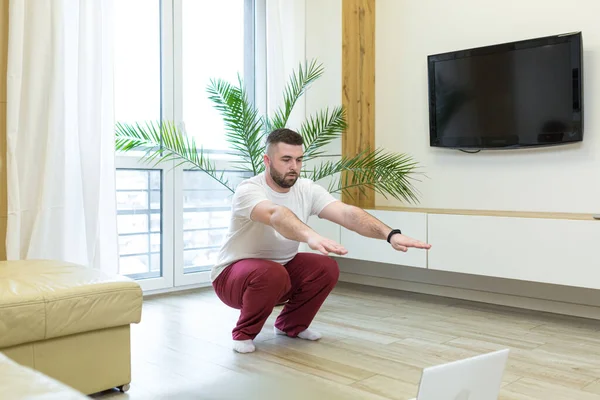 young adult bearded man doing stretching exercises at home in the living room watching video lesson or tutorial online using a laptop. Man doing fitness and sports in the morning sitting on the floor