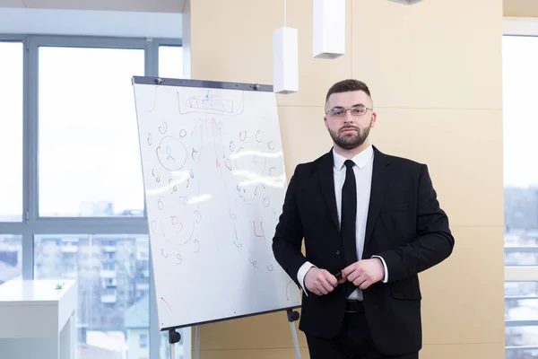Portrait young bearded man in a formal black business suit with glasses and tie looking at camera. Confident Male a modern office a whiteboard. Manager ceo meeting or conference person, businessman