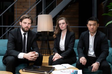 Çok ırklı iş adamları, erkekler ve kadınlar, bir iş toplantısında oturup kameraya bakıyorlar, iş dünyasından siyah takımlar giyiyorlar.