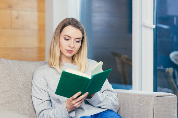 young woman sitting or lying on sofa at home and reading a book. student girl studying lessons. female relaxes indoors on a comfortable couch at leisure. nice to spend free time on the weekend