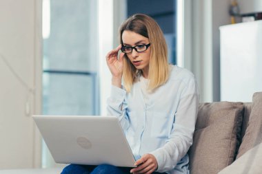 Evdeki kanepede günlük kıyafetlerle oturan güzel sarışın kadın ve arkadaşları ve ailesiyle video iletişimi için dizüstü bilgisayar kullanıyor.