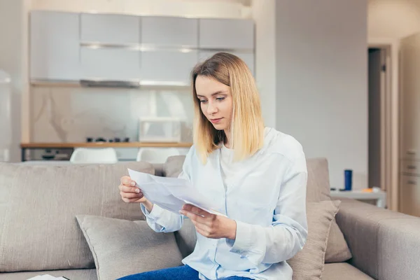 Happy blonde woman sitting at home on the couch, happy with timely payment of credit bills and closing the credit line, received good news from the bank