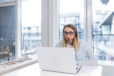 Kulaklık ve gözlüklü genç sarışın bir kadın müşterilerle çevrimiçi bir tüketici destek merkezinin ofisindeki bilgisayar ekranı ya da dizüstü bilgisayar ekranı önünde uzaktan iletişim kuruyor. konferans yardımı