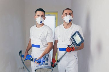 Sıvacı duvar yüzeyini bir duvar öğütücüsüyle düzleştiriyor. İki usta inşaatçı beyaz alçı bir duvarı öğütüyor. Tulumlu bir adam, yüzeyi solunum cihazıyla öğütür. deneyimli tamirci